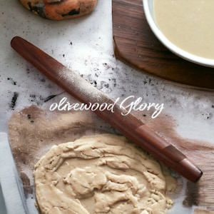May include: A dark brown wooden rolling pin, dusted with flour, lies on a surface with dough and the text "olivewood Glory". A bowl of batter and a wooden board are in the background. This is a kitchen tool for baking.