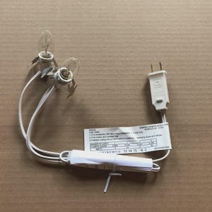 May include: A white electrical light cord set featuring two clear incandescent bulbs with metal clips, a two-prong plug, and a white informational tag from General Foam Plastics Corp. The tag lists 120V/60Hz specifications and safety instructions.