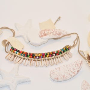 May include: A colourful beaded necklace with white shell accents. The necklace is made with a brown cord and has a variety of coloured beads, including red, orange, yellow, green, blue, and purple.