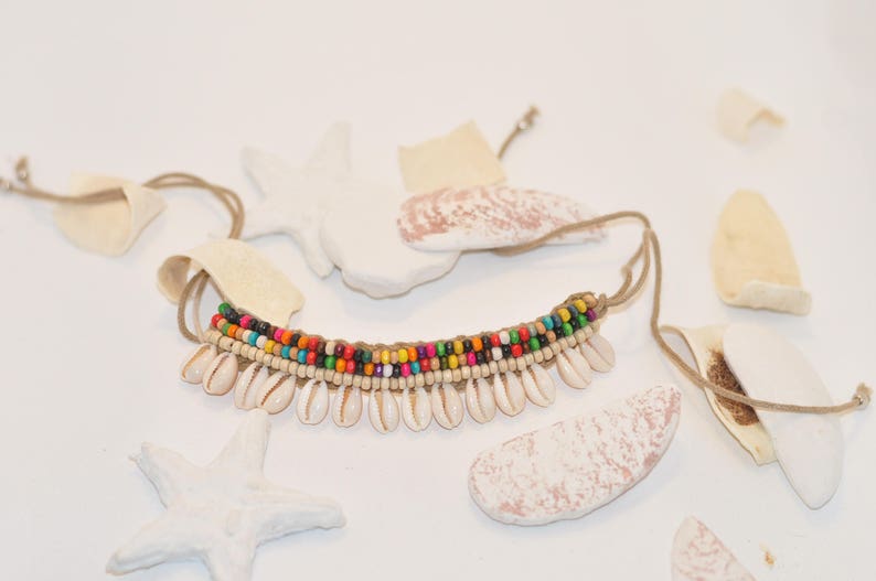 May include: A colourful beaded necklace with white shell accents. The necklace is made with a brown cord and has a variety of coloured beads, including red, orange, yellow, green, blue, and purple.