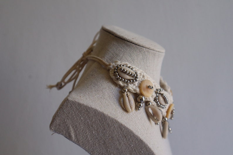 K&ouml;nnte beinhalten: Eine wei&szlig;e geh&auml;kelte Halskette mit Muscheln, Perlen und Kn&ouml;pfen. Die Halskette befindet sich auf einer wei&szlig;en Puppe.