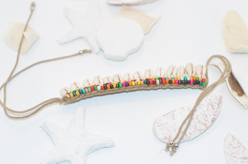 May include: A brown cord bracelet with white seashells and colourful beads. The bracelet is tied with a knot at the end.