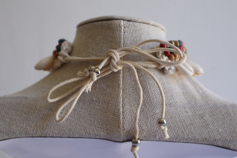 May include: A white and beige macrame necklace with colourful beads and shell accents. The necklace is tied with a simple knot and has silver metal beads on the ends of the cords.