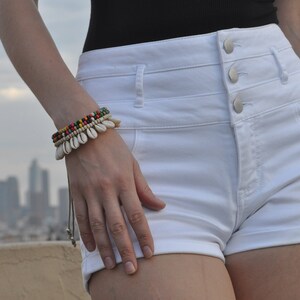 May include: White high-waisted denim shorts with a button fly and three buttons. The shorts have a rolled cuff and are being worn by a person with a bracelet on their wrist.