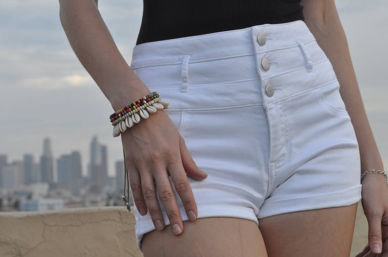 May include: White high-waisted denim shorts with a button fly and three buttons. The shorts have a rolled cuff and are being worn by a person with a bracelet on their wrist.