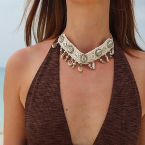 K&ouml;nnte beinhalten: Eine wei&szlig;e geh&auml;kelte Choker-Halskette mit silbernen Akzenten und Muschel-Anh&auml;ngern. Die Kette wird von einer Person mit braunen Haaren und einem braunen Oberteil getragen.