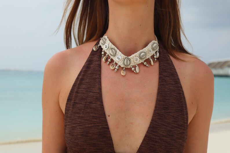 K&ouml;nnte beinhalten: Eine wei&szlig;e geh&auml;kelte Choker-Halskette mit silbernen Akzenten und Muschel-Anh&auml;ngern. Die Kette wird von einer Person mit braunen Haaren und einem braunen Oberteil getragen.