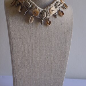 K&ouml;nnte beinhalten: Eine wei&szlig;e geh&auml;kelte Choker-Kette mit silbernen Perlen, Muschelakzenten und wei&szlig;en Kn&ouml;pfen. Die Kette wird auf einer stoffbezogenen Puppe pr&auml;sentiert.