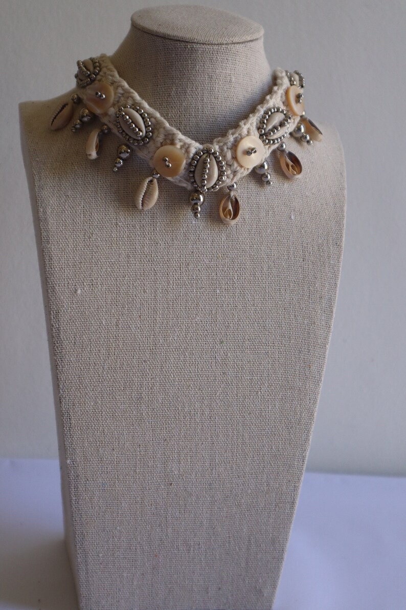 K&ouml;nnte beinhalten: Eine wei&szlig;e geh&auml;kelte Choker-Kette mit silbernen Perlen, Muschelakzenten und wei&szlig;en Kn&ouml;pfen. Die Kette wird auf einer stoffbezogenen Puppe pr&auml;sentiert.