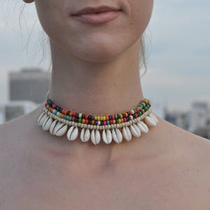 May include: A multi-coloured beaded choker necklace with white shell accents. The necklace is made of small, colourful beads and has a row of white shells hanging from it.