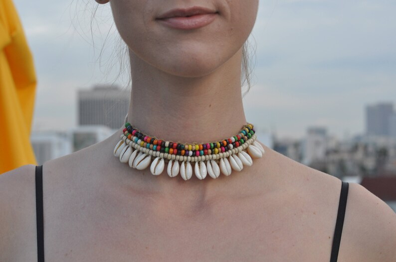 May include: A multi-coloured beaded choker necklace with white shell accents. The necklace is made of small, colourful beads and has a row of white shells hanging from it.