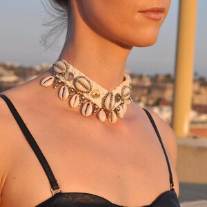 K&ouml;nnte beinhalten: Eine wei&szlig;e geh&auml;kelte Choker-Halskette mit Muschelakzenten. Die Halskette ist mit kleinen wei&szlig;en Muscheln und silbernen Perlen verziert.