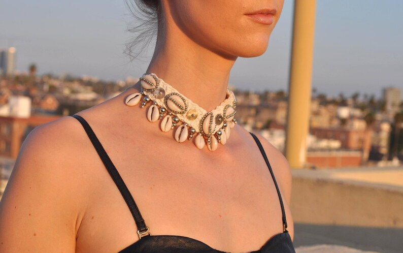 K&ouml;nnte beinhalten: Eine wei&szlig;e geh&auml;kelte Choker-Halskette mit Muschelakzenten. Die Halskette ist mit kleinen wei&szlig;en Muscheln und silbernen Perlen verziert.
