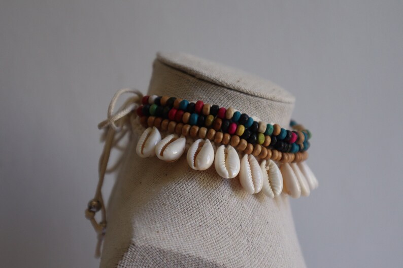 May include: A multi-strand anklet with wooden beads in brown, black, blue, green, red, and pink. The anklet is adorned with white seashells.