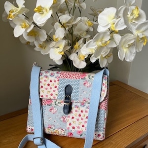 May include: A floral patchwork shoulder bag with a light blue strap and buckle closure. The bag features a mix of floral patterns in pink, red, and blue. The bag is sitting on a wooden surface, with white orchids in the background.
