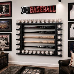 May include: A baseball-themed room with framed sports memorabilia. A black shelf displays baseball bats and baseballs in protective cases. A neon sign reads "BASEBALL." Framed jerseys and a Babe Ruth poster are also visible.