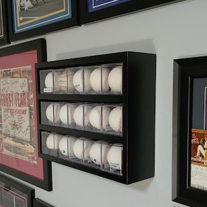 May include: A black baseball display case with three rows of clear plastic compartments, each holding a white baseball. The case is mounted on a wall.