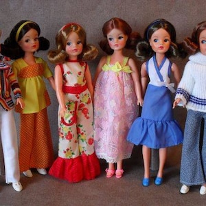 Op de afbeelding: Zeven vintage poppen met lang haar die verschillende outfits dragen. De poppen dragen een verscheidenheid aan kleuren en patronen, waaronder strepen, stippen en bloemenprints. De poppen staan ​​allemaal op een lichtbruin oppervlak.