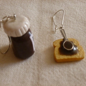 Peut inclure: Une paire de boucles d'oreilles fantaisie. L'une représente un petit pot de pâte à tartiner au chocolat avec un couvercle blanc. L'autre est une tranche de pain grillé avec de la pâte à tartiner et une petite cuillère.