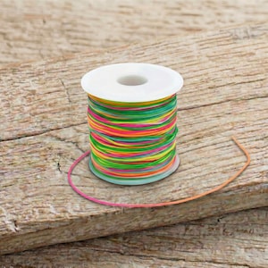 Peut inclure: Une bobine de fil élastique multicolore, avec des teintes vives de rose, jaune, vert et orange. Le fil est enroulé autour d'une bobine en plastique blanc, parfait pour la création de bijoux ou les travaux manuels.