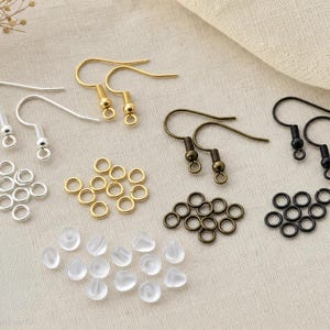 May include: A selection of earring-making components. The image shows silver, gold, bronze, and black earring hooks, jump rings, and clear earring backs. The items are arranged on a neutral fabric.