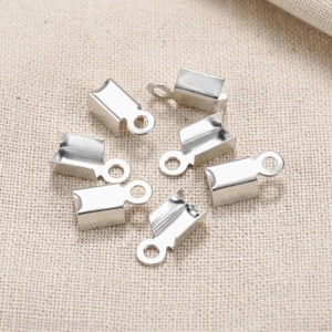 May include: Six silver-colored crimp end findings for jewelry making. Each finding has a rectangular shape with a loop at one end for attaching to a chain or other components. The findings are scattered on a neutral-toned fabric.