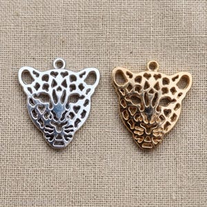 Puede incluir: Dos colgantes metálicos con forma de cabeza de leopardo. Uno es plateado y el otro dorado. Ambos colgantes tienen un diseño recortado y un pequeño lazo en la parte superior para sujetarlos a una cadena u otro componente de joyería. Sobre un fondo de tela beige.