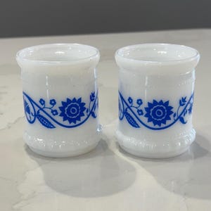 May include: Two white milk glass cups with blue floral designs. The cups are approximately 3 inches tall and 2 inches in diameter.