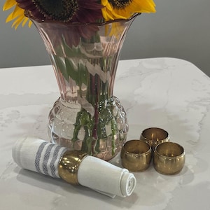 May include: Three gold napkin rings on a white countertop next to a pink glass vase filled with sunflowers and burgundy flowers.