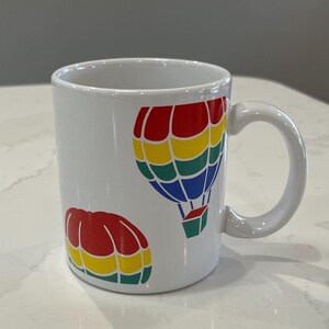 May include: White ceramic mug with a red, yellow, blue, and green hot air balloon design. The mug has a handle and is decorated with two hot air balloons.