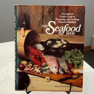 May include: A vintage cookbook titled "The Seafood Book: A Complete Cook's Guide to Preparing and Cooking Fish and Shellfish" by Shirley Ross. The cover features a colorful illustration of fresh seafood, including lobster, fish, and clams.