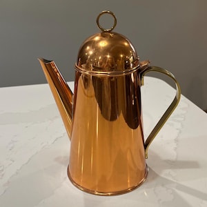 May include: A copper teapot with a brass handle and lid. The teapot has a shiny, polished finish.
