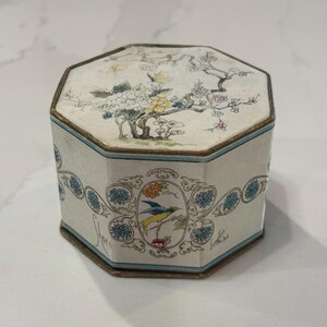 May include: An octagonal box with a cream-colored base featuring a lid decorated with a floral and butterfly illustration. The sides display a repeating pattern of blue flowers and a central oval medallion containing a bird, with the name Shari inscribed.