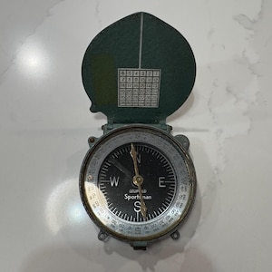 May include: A vintage Leupold Sportsman compass with a dark green cover and a black face. The compass features directional markings, a rotating bezel, and a needle pointing south. The cover has a grid of numbers.