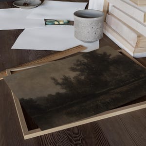 May include: A framed landscape print depicting a dark, atmospheric scene of trees reflected in water. The print is in a light wooden frame, on a wooden surface with art supplies and a ceramic cup.