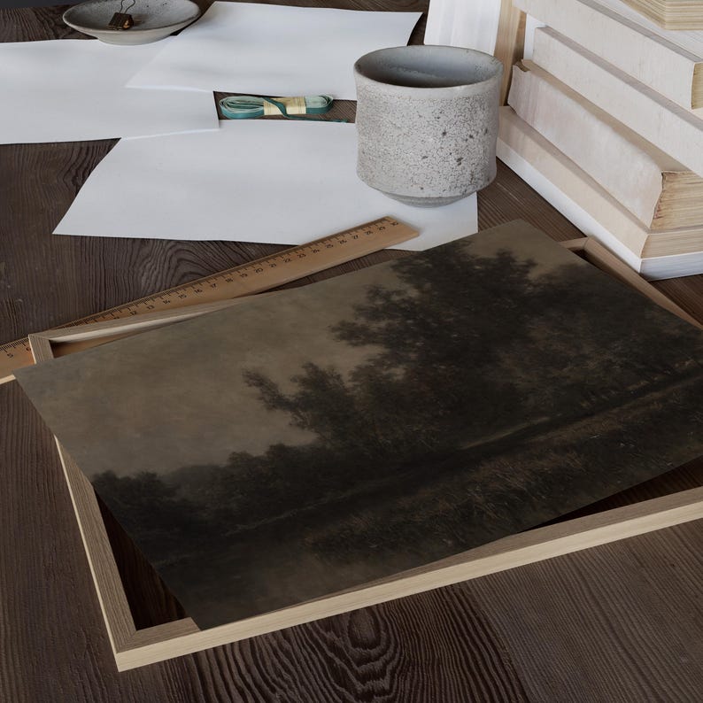 May include: A framed landscape print depicting a dark, atmospheric scene of trees reflected in water. The print is in a light wooden frame, on a wooden surface with art supplies and a ceramic cup.