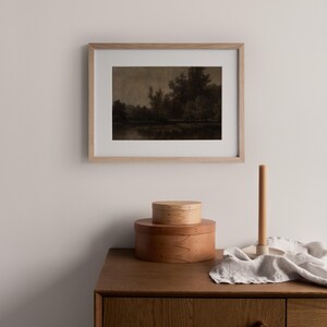 May include: A framed sepia-toned landscape print hangs above a wooden cabinet. Two round wooden boxes are stacked on the cabinet, with a candle and holder to the right. The print depicts trees and water.