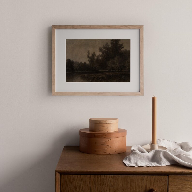 May include: A framed sepia-toned landscape print hangs above a wooden cabinet. Two round wooden boxes are stacked on the cabinet, with a candle and holder to the right. The print depicts trees and water.