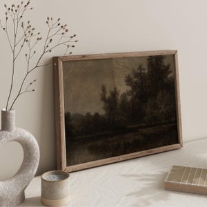 May include: A framed landscape painting in sepia tones. The artwork shows a dark, atmospheric scene of trees and water. The frame is light-coloured wood. A decorative vase, candle, and small wooden box are in the foreground.
