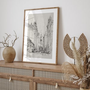 May include: A black and white framed print of a city street scene with tall buildings. The print is hanging on a wall above a wooden cabinet with a woven rattan front.