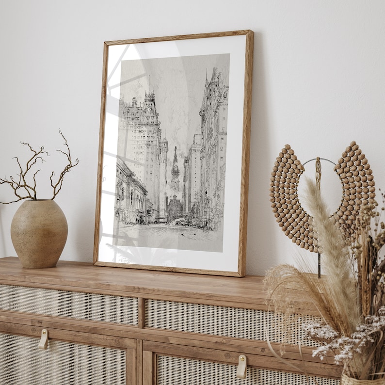 May include: A black and white framed print of a city street scene with tall buildings. The print is hanging on a wall above a wooden cabinet with a woven rattan front.