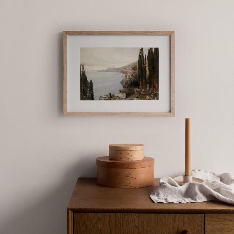 May include: A framed print of a seascape painting with a view of a coastal town and a body of water. The painting is in a light wood frame and is hanging on a white wall.