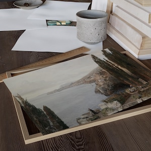 May include: A framed watercolor painting depicting a coastal scene with a town nestled on a hillside overlooking a calm blue sea. Lush green trees line the hillside and the water's edge.