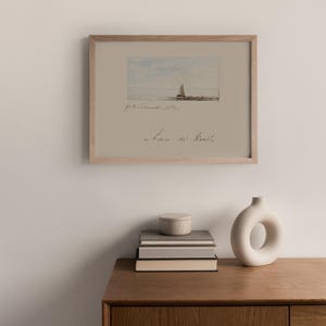 Puede incluir: Una impresi&oacute;n en acuarela enmarcada de un velero en el mar, con el texto "Jan de Kust" y otras escrituras. La impresi&oacute;n est&aacute; en un marco de madera clara y se exhibe sobre una superficie de madera con una pila de libros, un peque&ntilde;o recipiente redondo y un jarr&oacute;n blanco.