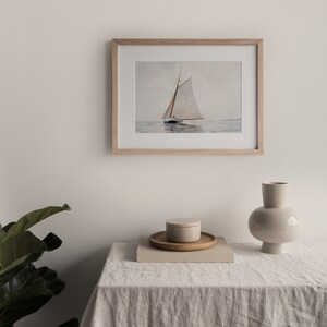 Puede incluir: Una pintura de acuarela enmarcada de un velero con velas blancas en un cuerpo de agua tranquilo. La pintura est&aacute; en un marco de madera clara y est&aacute; colgada en una pared blanca.