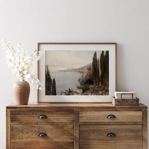 May include: A framed landscape painting depicting a coastal scene with a view of a town and a body of water. The painting is in a brown frame and is hanging on a wall.