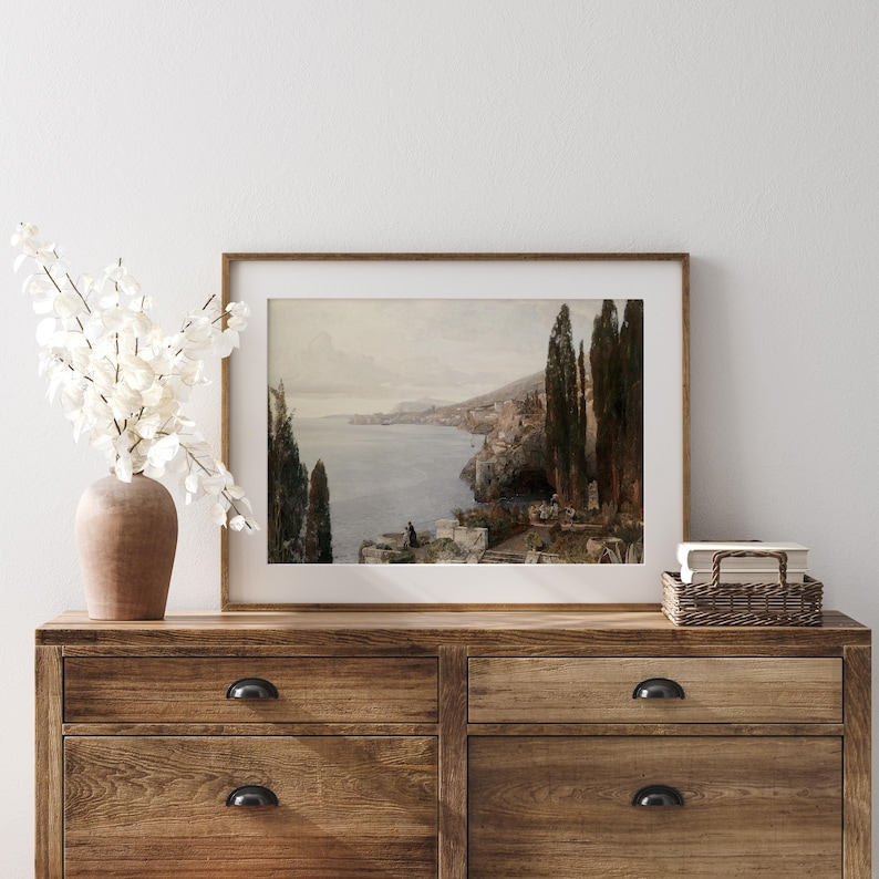 May include: A framed landscape painting depicting a coastal scene with a view of a town and a body of water. The painting is in a brown frame and is hanging on a wall.