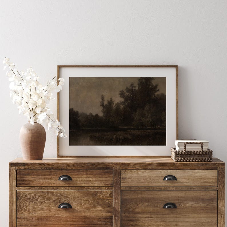May include: A framed landscape print depicting a dark, atmospheric scene of trees and water. The artwork is displayed on a wooden chest of drawers, alongside a vase of white flowers and a small wicker basket.