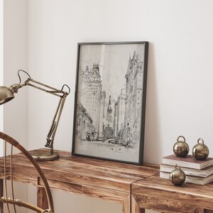 May include: A black and white framed print of a city street scene with tall buildings. The print is hanging on a white wall above a wooden table with a lamp and decorative ornaments.