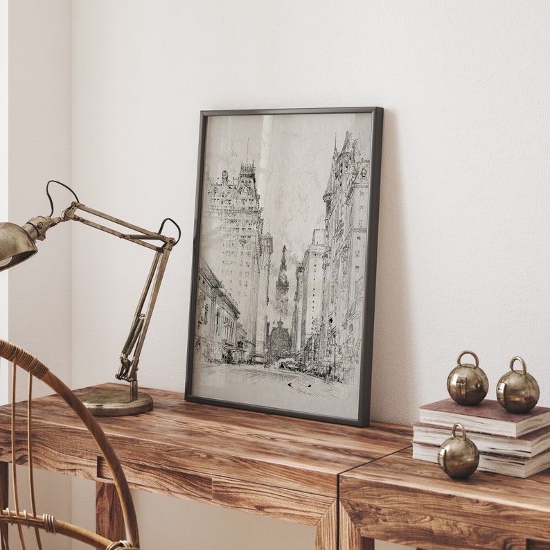 May include: A black and white framed print of a city street scene with tall buildings. The print is hanging on a white wall above a wooden table with a lamp and decorative ornaments.